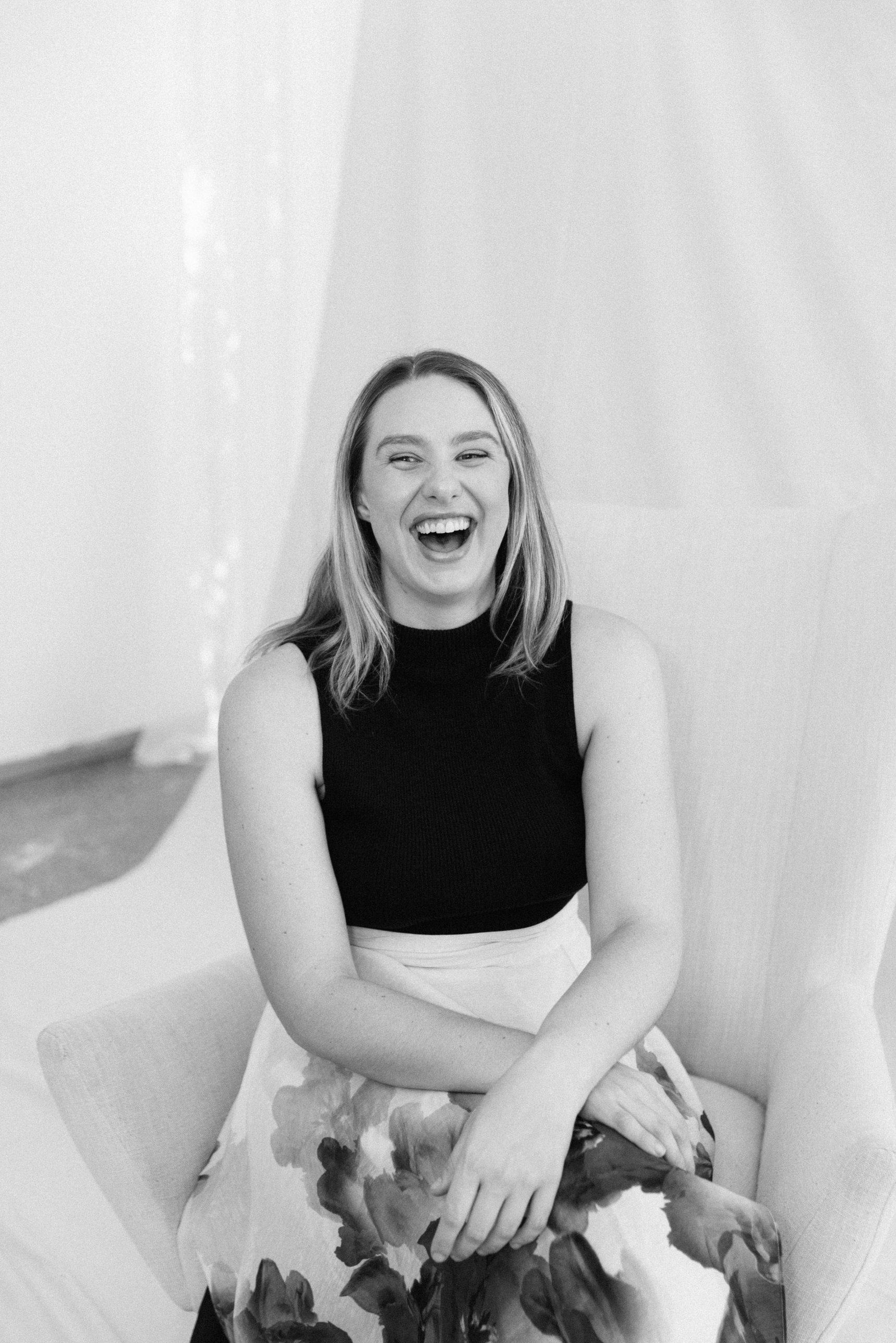 Black and white portrait of Ashleigh Kay laughing, seated in an armchair
